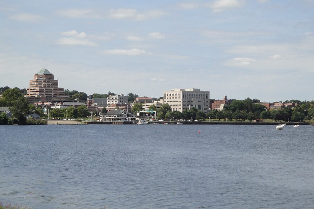 Downtown Middletown seen across the wide Connecticut River, with a tiered tower topped by a green pyramid roof, an office block and small boats near the shore