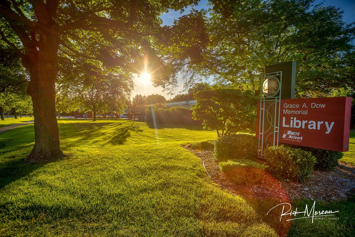 The red Grace A. Dow Memorial Library sign on a green lawn, with the summer sun shining through large shade trees