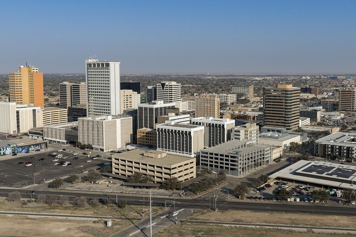Downtown Midland seen from above on a clear day, office towers and a parking garage beside a large surface lot, with a rail line and highway in the foreground
