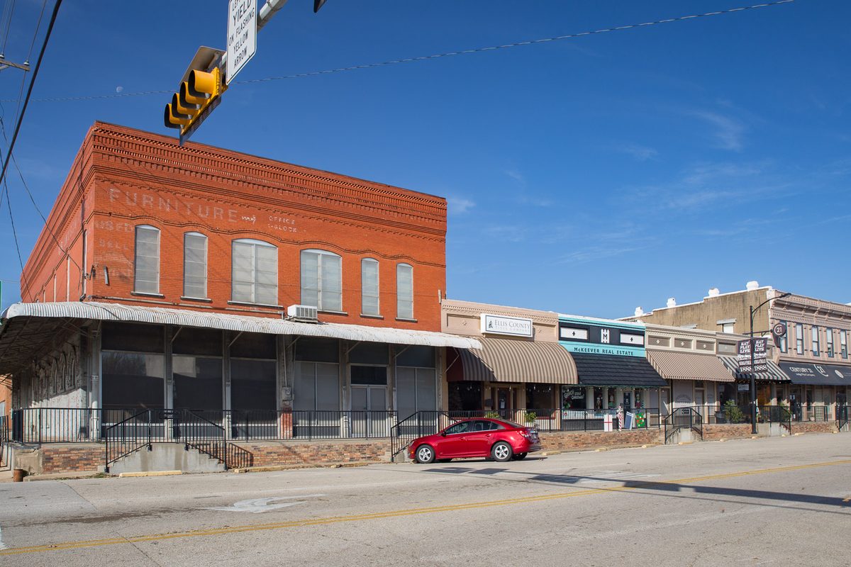 A red-brick former furniture store on a downtown Midlothian corner beside a row of awning-covered shops, a red car parked at the curb under a traffic light