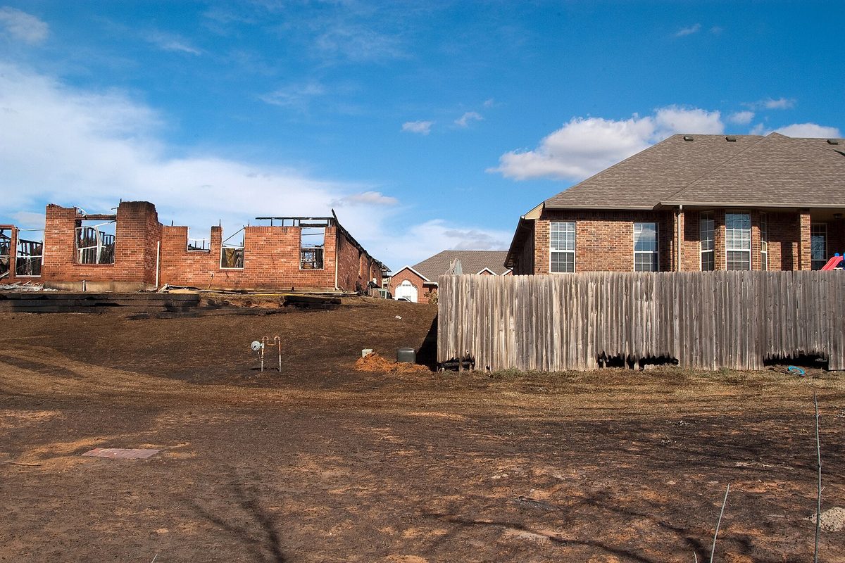 The burned-out brick shell of a house beside an undamaged brick home and wooden fence in Midwest City after the March 2009 wildfires, scorched ground in front