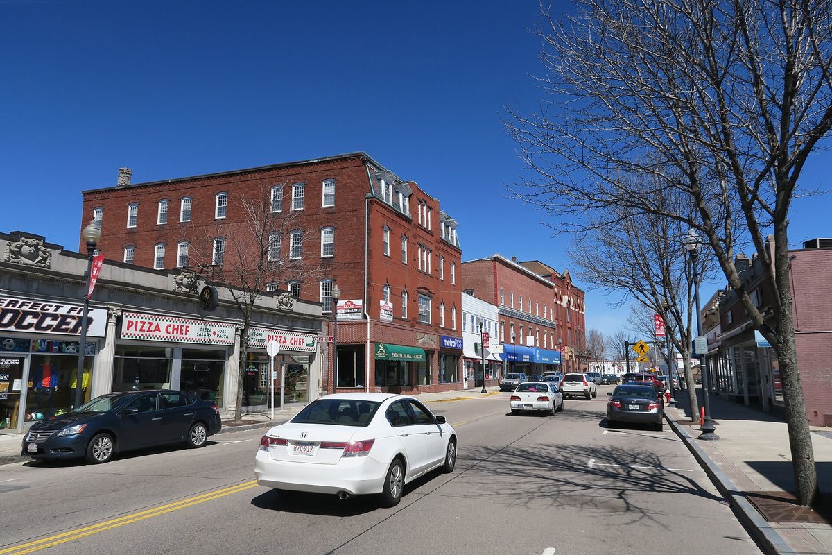 Main Street in Milford under a blue sky, with red brick storefronts, a pizza shop and a white sedan driving past bare trees