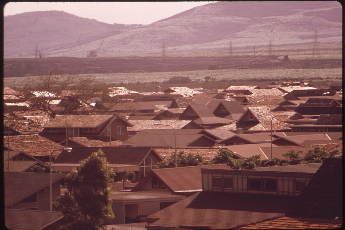An old color photograph of rows of brown-roofed tract houses, with dry hills and power-line towers in the hazy distance