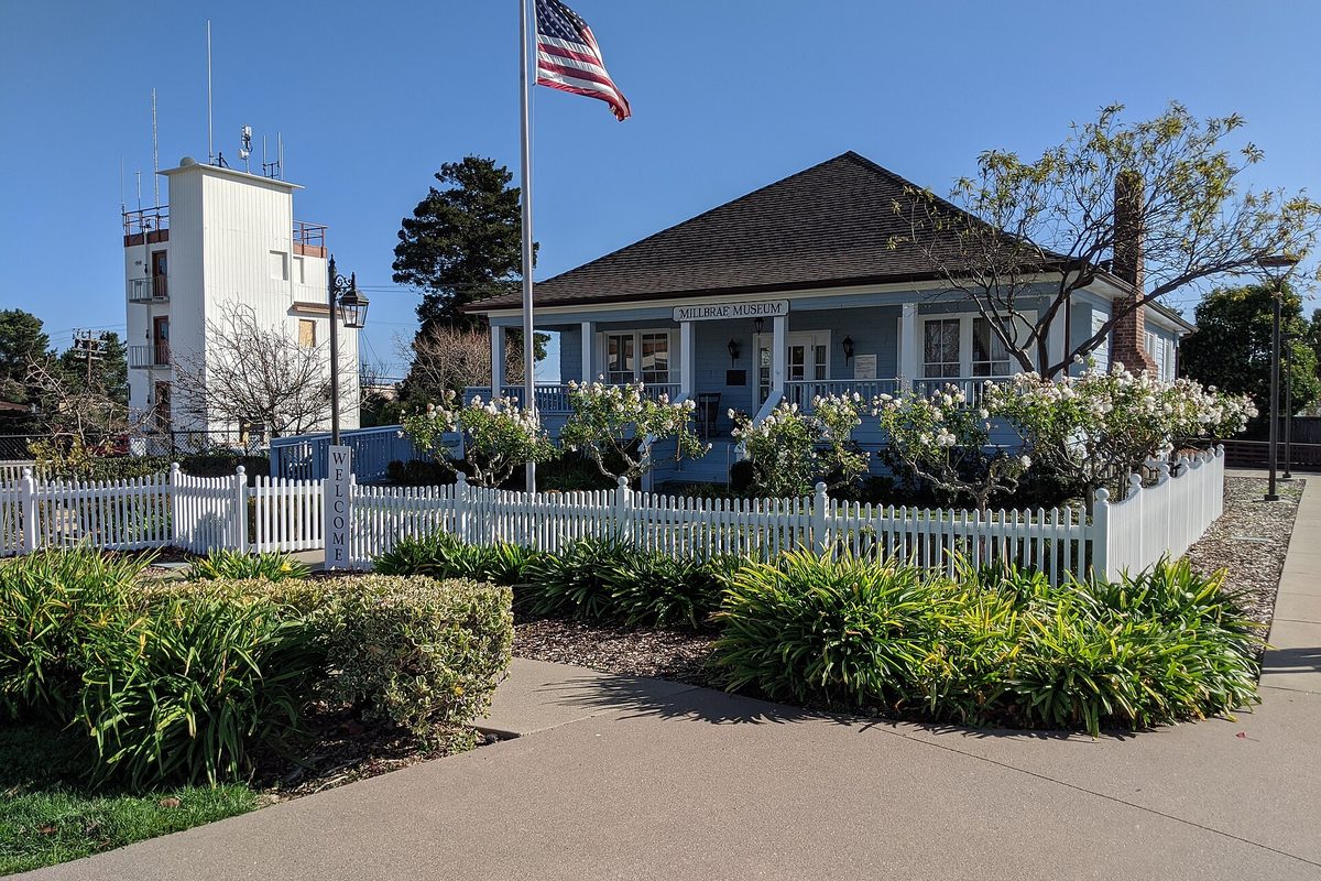 The Millbrae Museum, a blue single-story house with a white picket fence and an American flag, beside a white tower