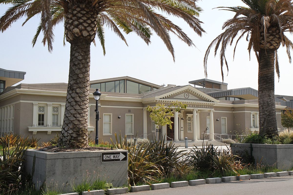 The old Milpitas Grammar School, a tan building with white columns, framed by two tall palm trees and a one-way sign