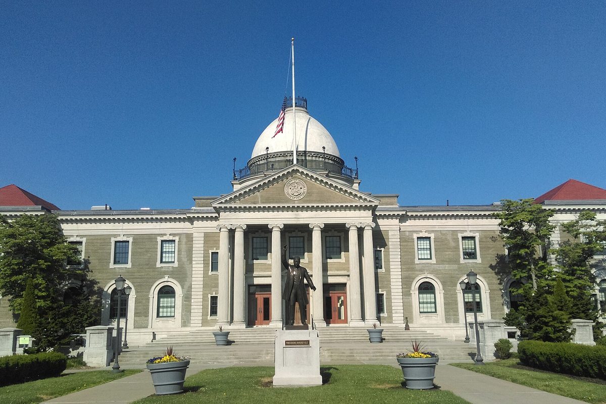 The domed, columned county building at 1550 Franklin Avenue in Mineola, with a bronze statue on a pedestal in front of its steps under a clear blue sky