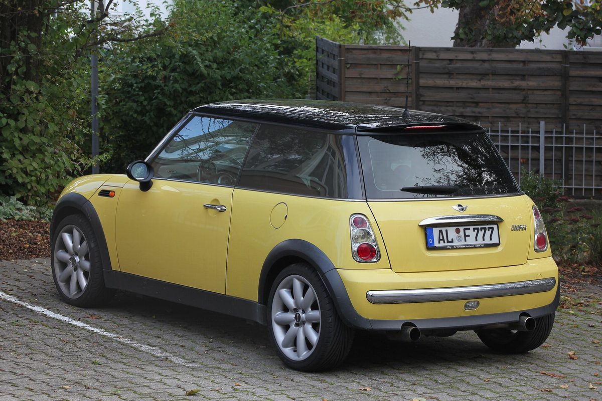 A yellow first-generation MINI Cooper hardtop with a black roof and silver wheels, seen from the rear on a cobblestone lot