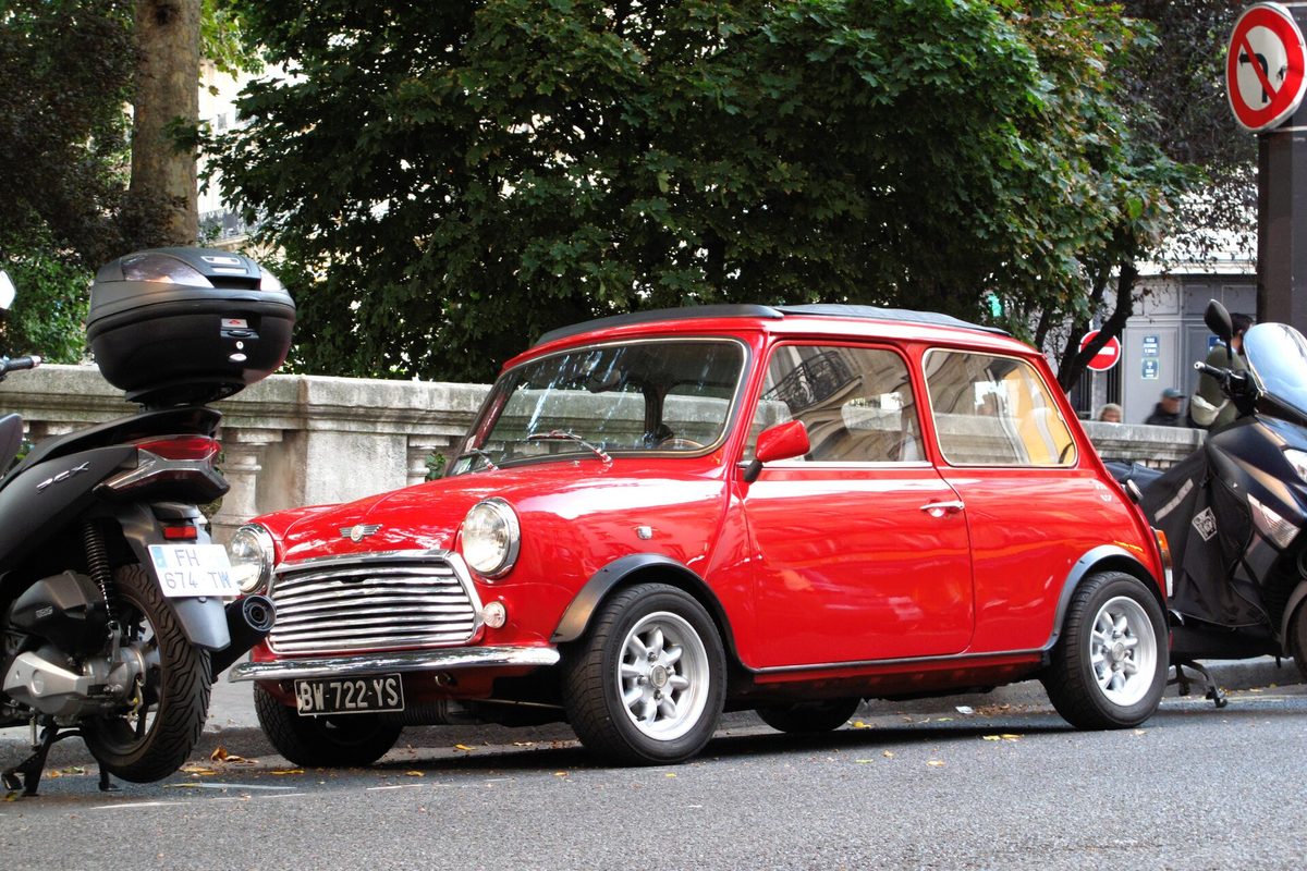 A small red classic Mini Cooper parked at the curb beside scooters on a city street