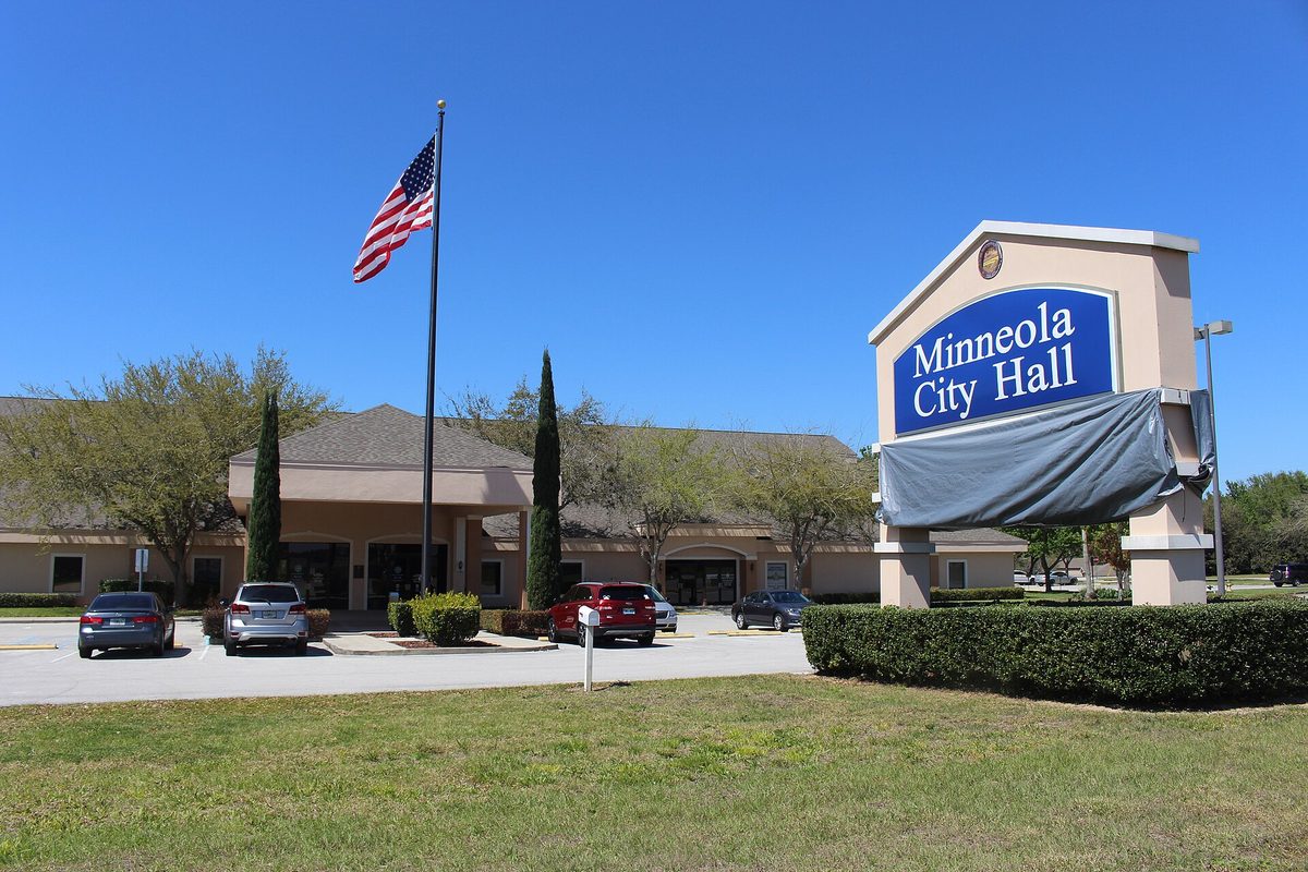The Minneola City Hall sign beside a flagpole and a low tan building with cars parked out front, under a clear blue sky