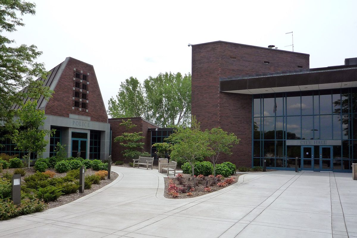 Minnetonka's brick City Hall with a tall glass entrance and the Police building beside it, joined by a curving concrete walkway lined with shrubs and flower beds