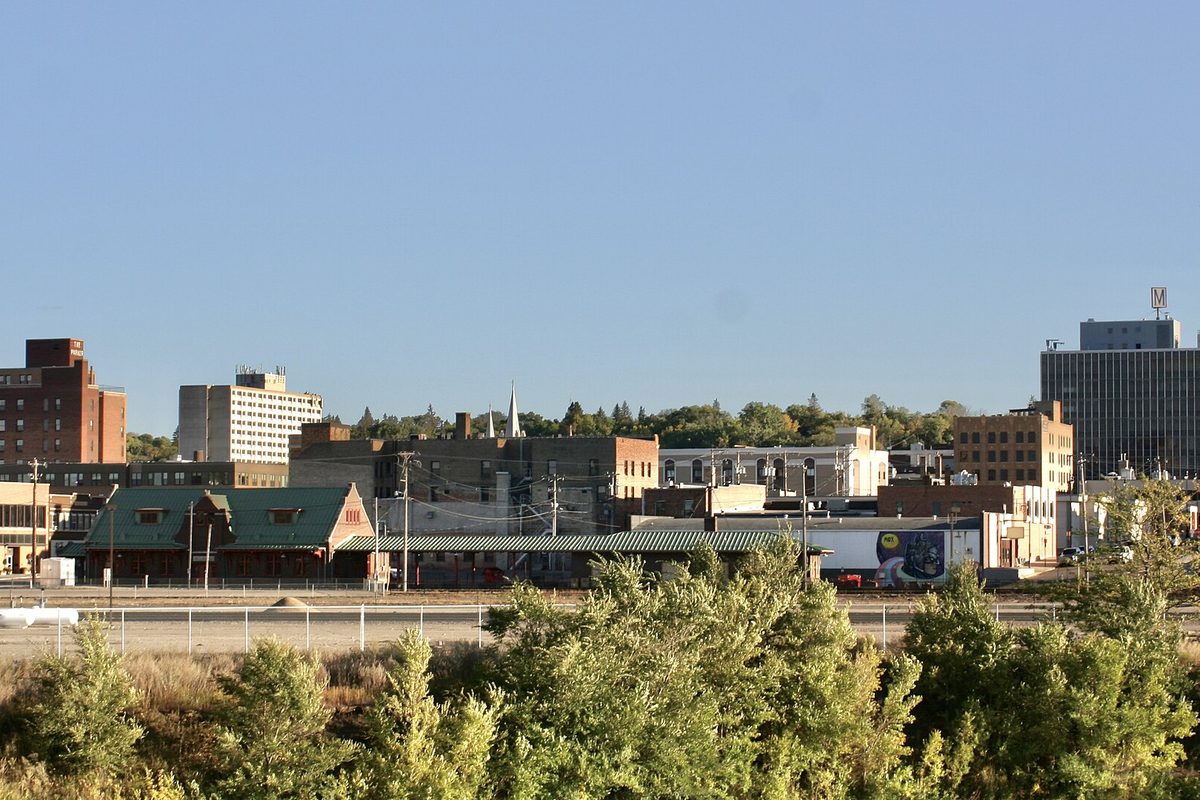 Downtown Minot seen across a rail yard, with a green-roofed depot, brick buildings and a church steeple above a line of green shrubs