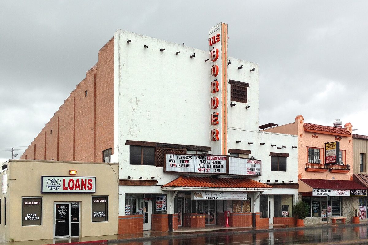 The Border Theatre in Mission, a white stucco building with a tall vertical red-lettered sign and a marquee, beside a loan office on a wet street