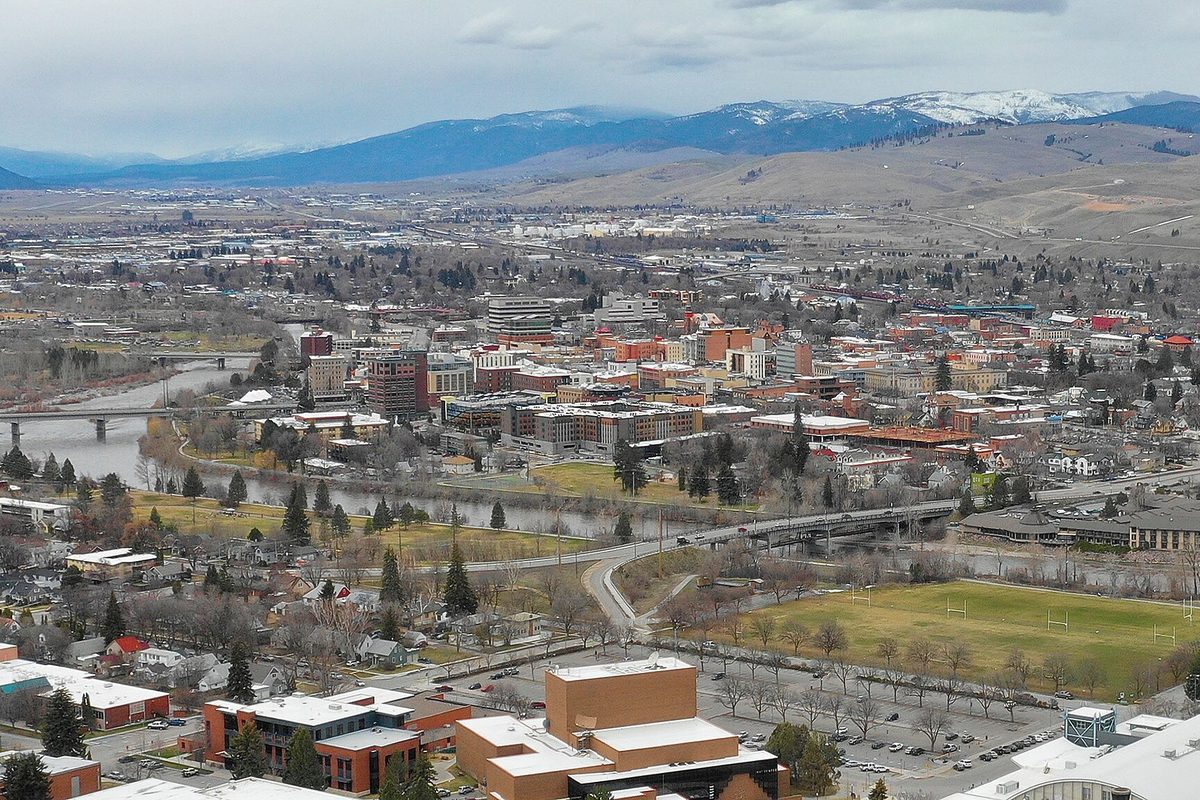 Downtown Missoula's brick buildings beside a river crossed by bridges, with playing fields in front and snow-topped mountains under a gray sky