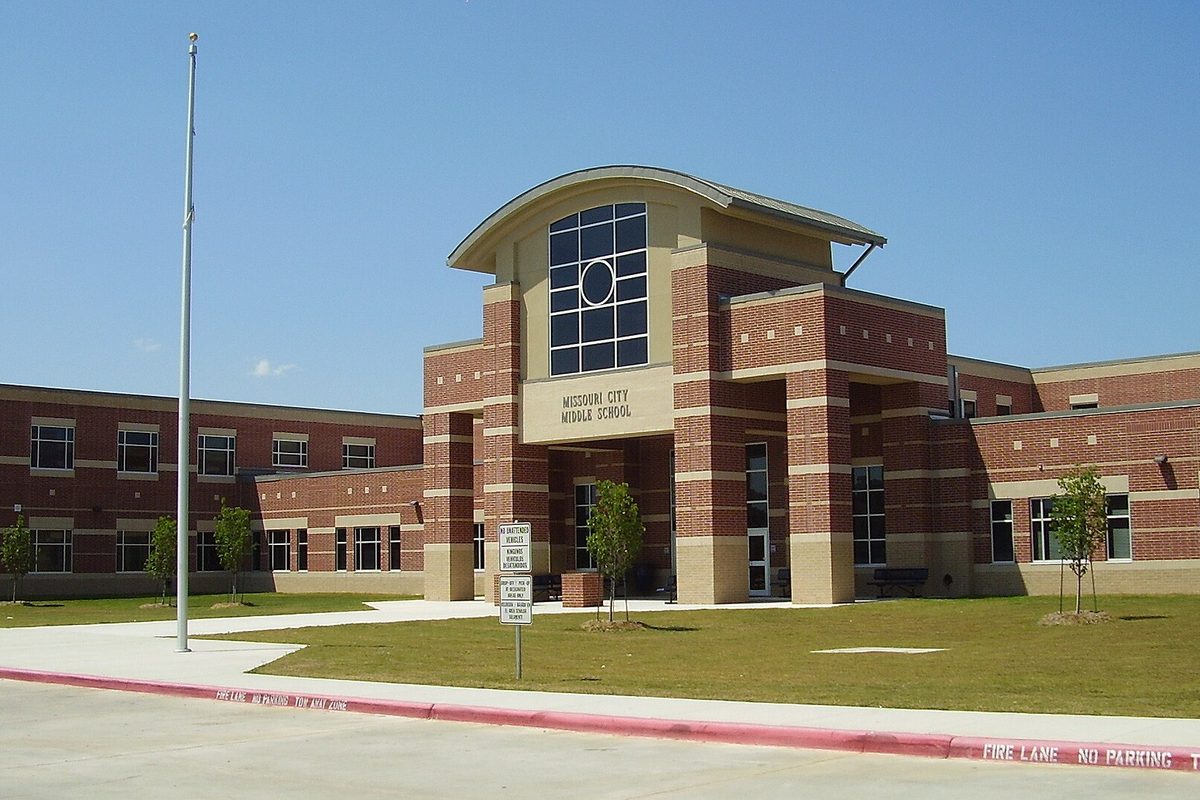 Brick front entrance of Missouri City Middle School with a tall arched window, a flagpole, young trees and a red-curbed fire lane