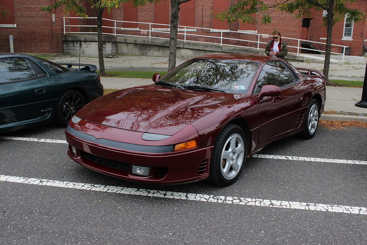 A burgundy Mitsubishi 3000GT liftback coupe with a rear spoiler and silver wheels, parked in a lined lot beside a dark green car