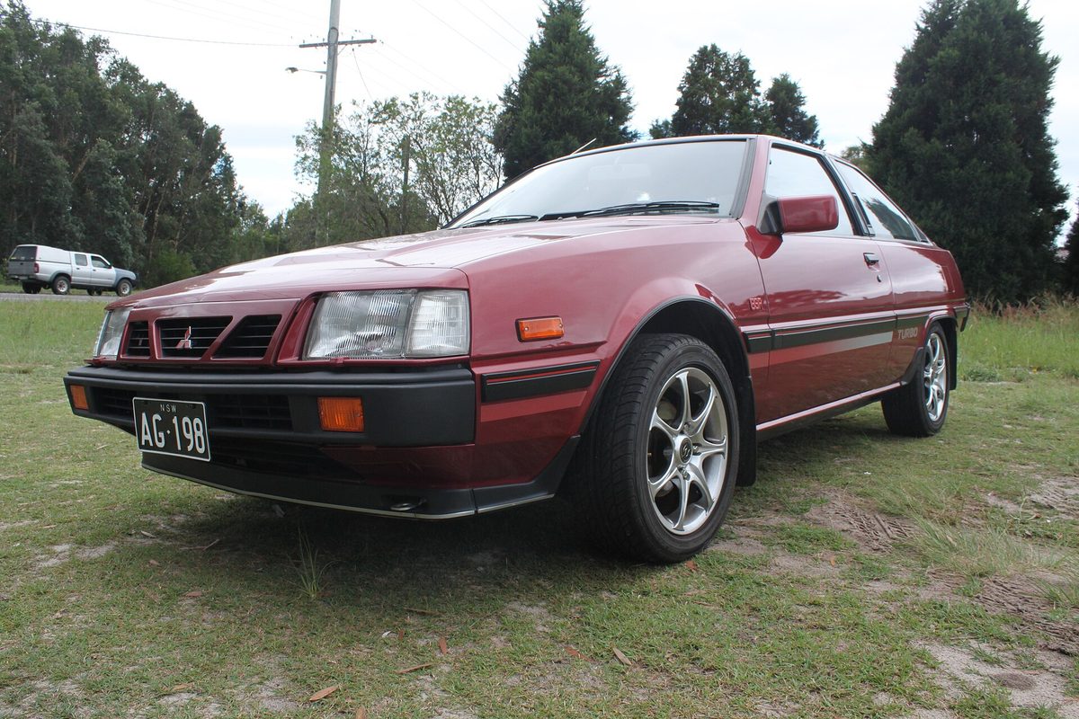 A maroon Mitsubishi Cordia hatchback with black lower trim and alloy wheels, parked on grass under a cloudy sky