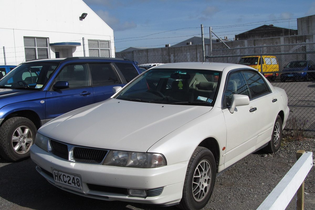 A white Mitsubishi Diamante sedan with silver alloy wheels parked on gravel beside a blue SUV and a chain-link fence