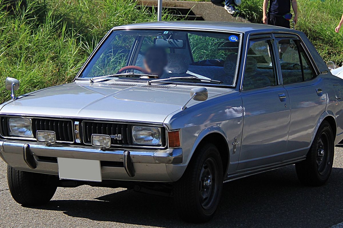 A silver 1969 Mitsubishi Colt Galant four-door sedan with chrome bumpers, driving past a grassy bank