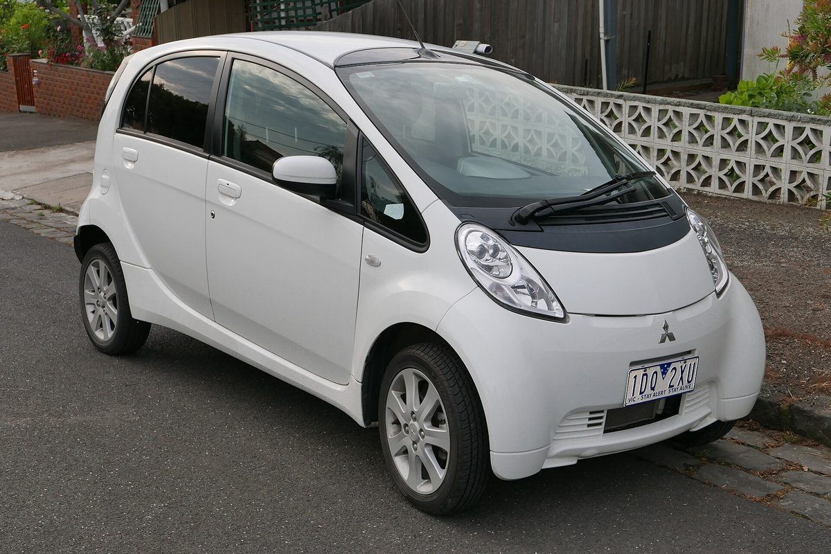 A white Mitsubishi i-MiEV small electric hatchback parked at a curb in front of a brick and concrete-block wall