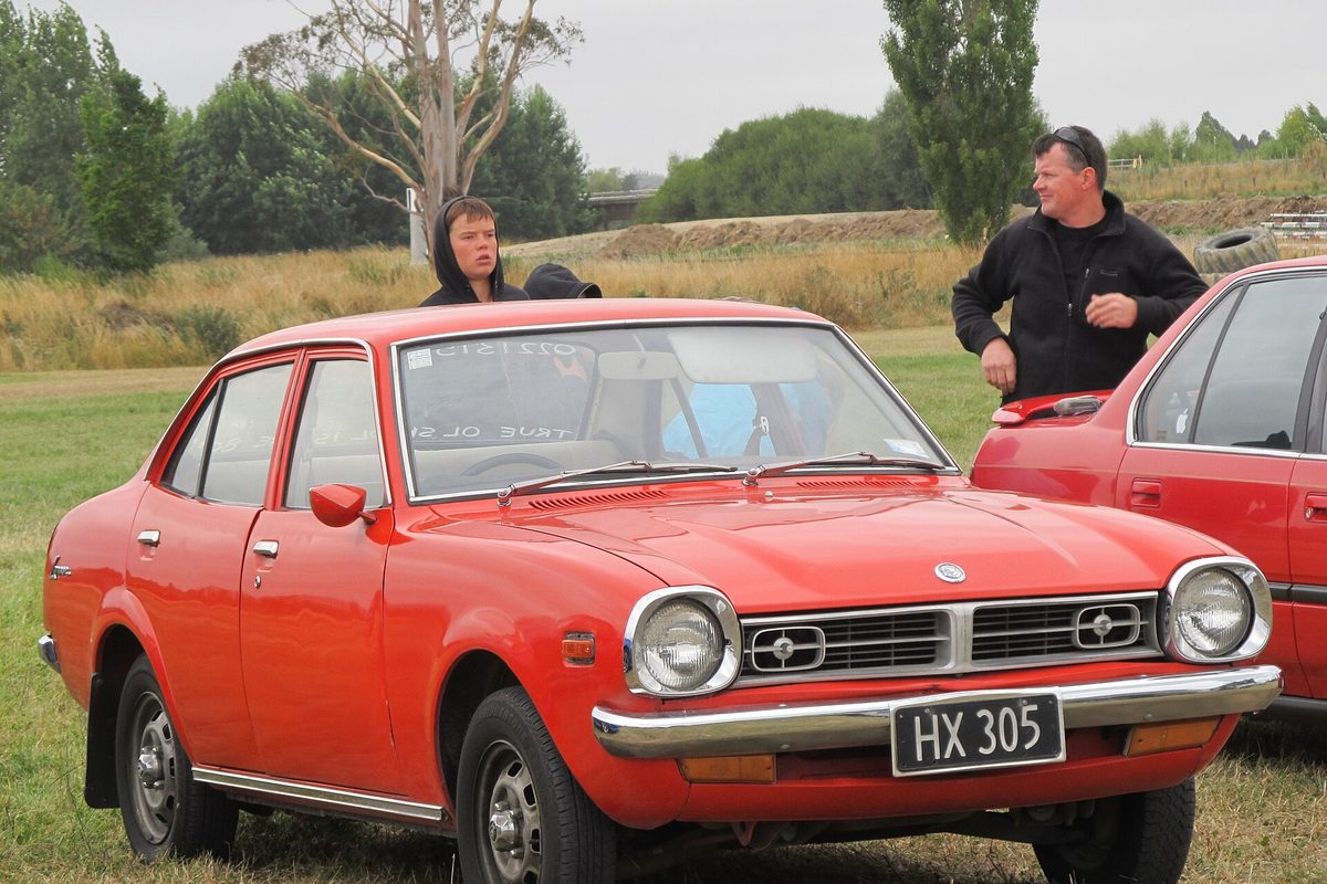 A red 1976 Mitsubishi Lancer four-door sedan on the grass at a car show, with two men standing behind it