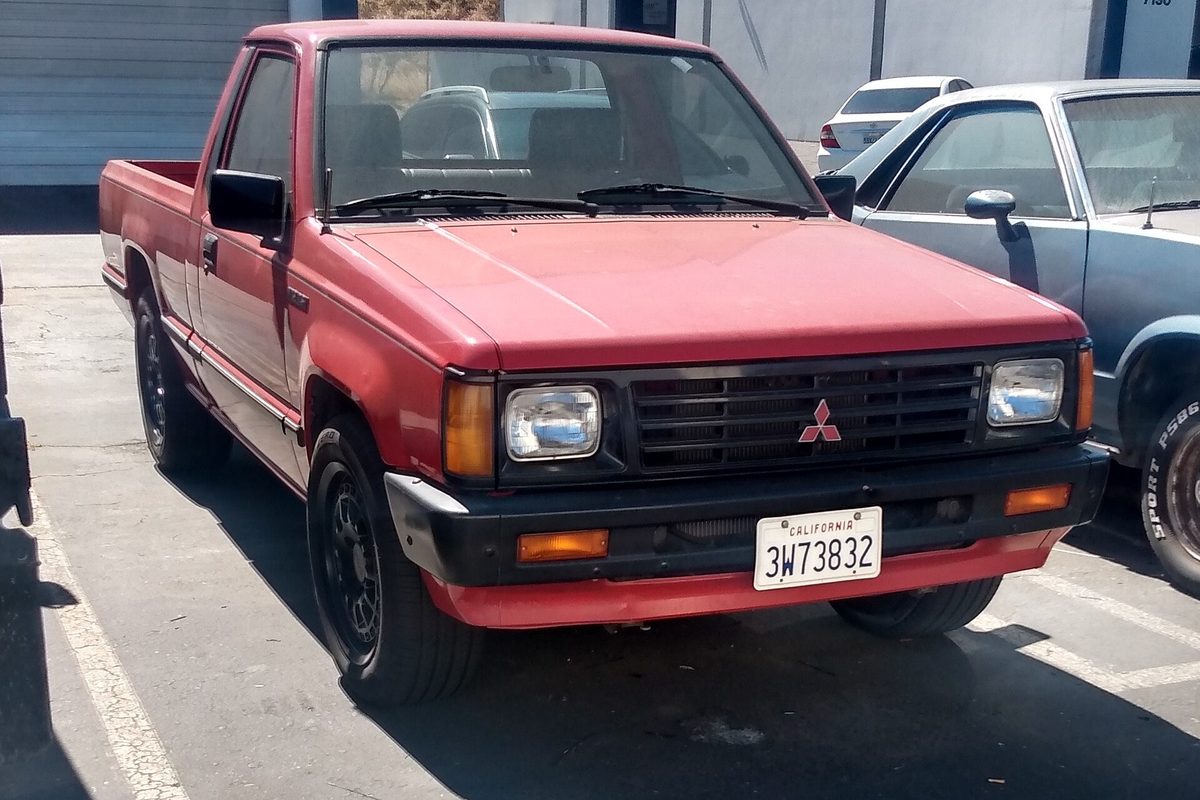 A red regular-cab Mitsubishi Mighty Max pickup with a black front bumper, parked in a sunny lot next to a gray sedan