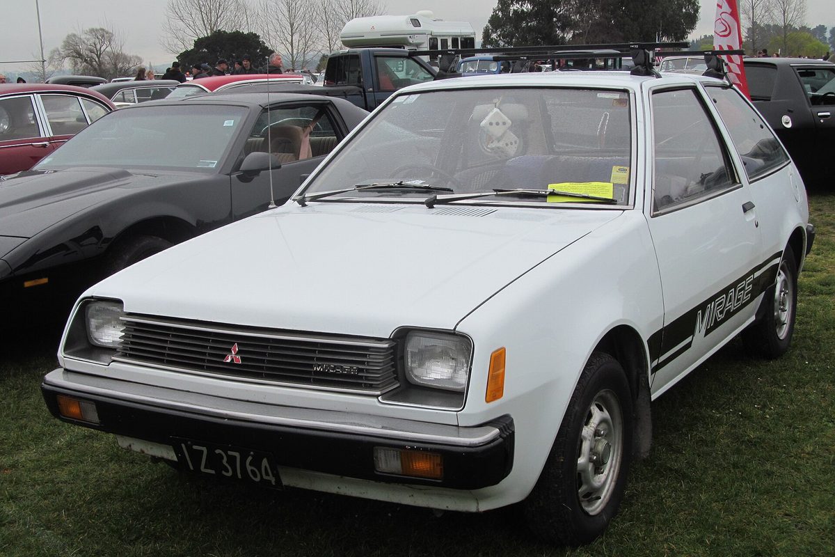 A white 1979 Mitsubishi Mirage three-door hatchback with a black Mirage side stripe, parked on grass at a car show
