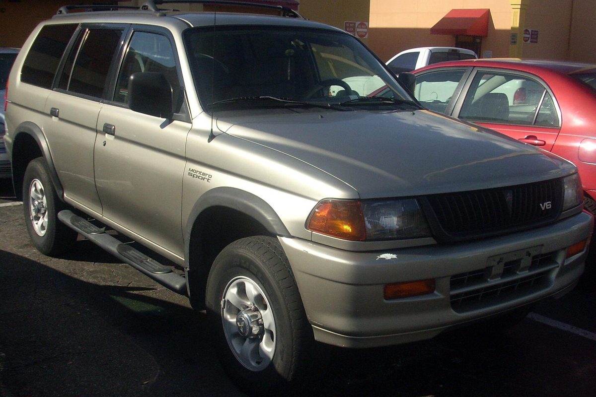 A champagne Mitsubishi Montero Sport SUV with side steps and a V6 badge on the grille, parked next to a red car