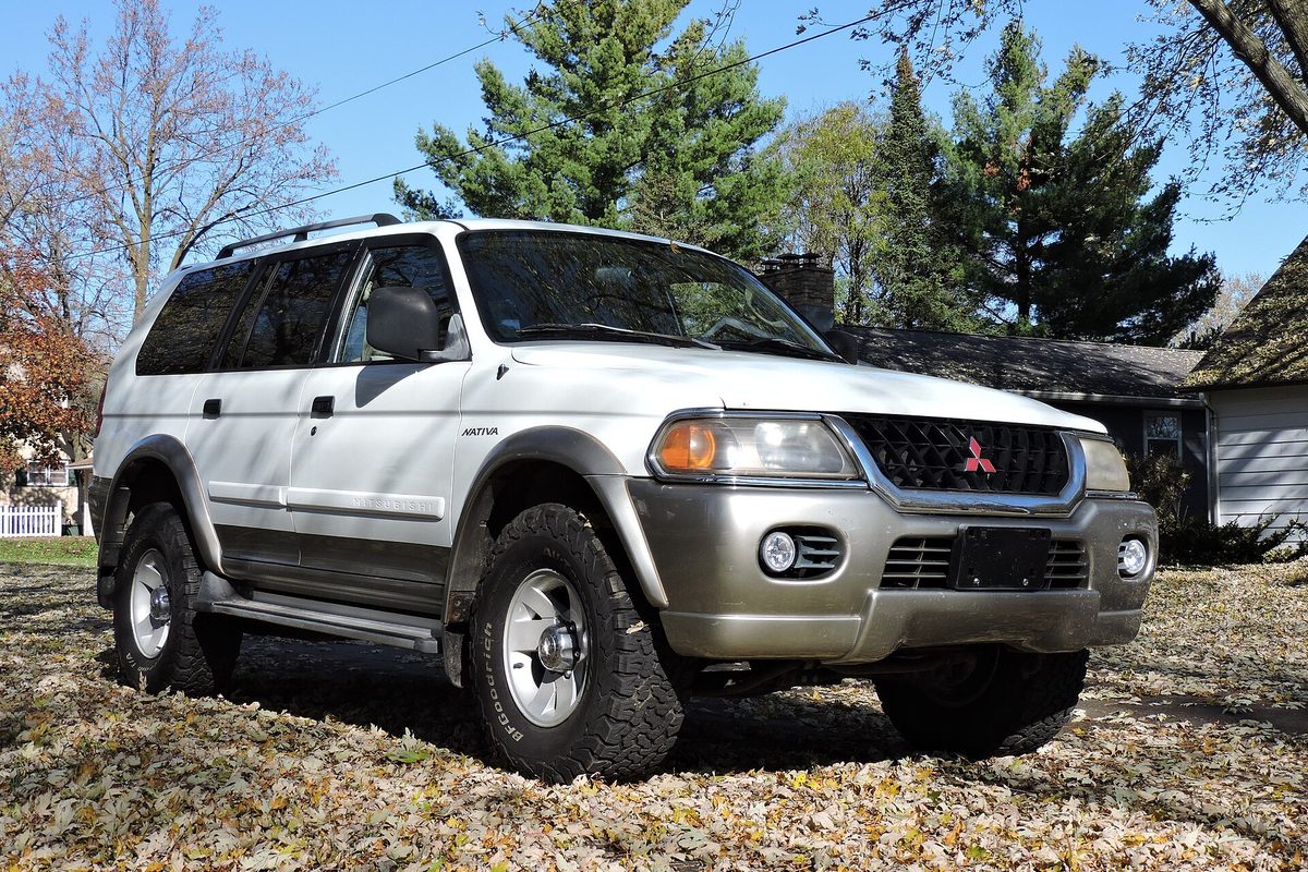 A white Mitsubishi Nativa SUV with champagne lower trim and a roof rack, parked on fallen leaves