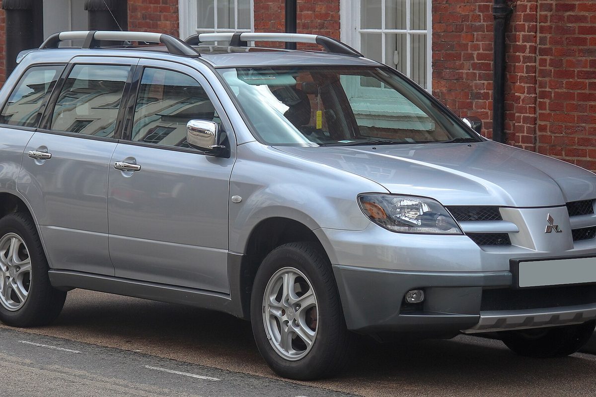 A silver first-generation Mitsubishi Outlander with roof rails, parked at a curb in front of a brick building