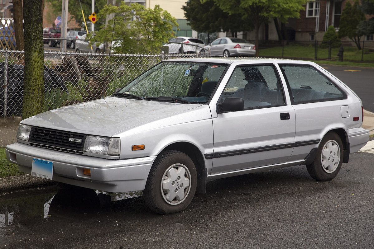 A silver Mitsubishi Precis three-door hatchback parked on a wet street beside a chain-link fence
