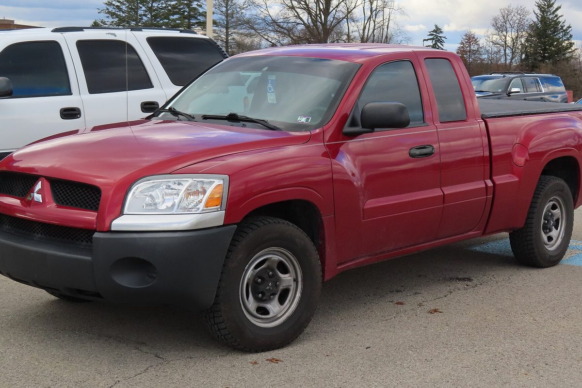 A red Mitsubishi Raider extended-cab pickup with steel wheels, parked in a lot next to a white SUV
