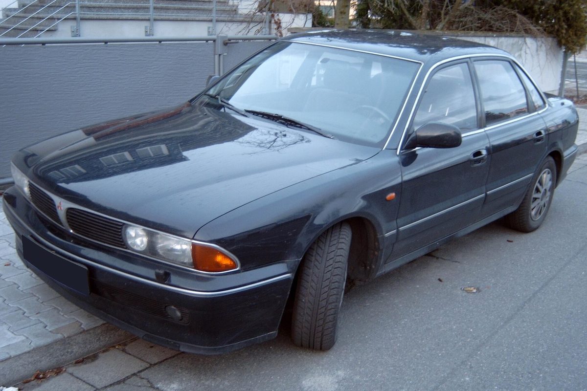 A dark navy Mitsubishi Sigma four-door sedan parked on a paved sidewalk beside a wall