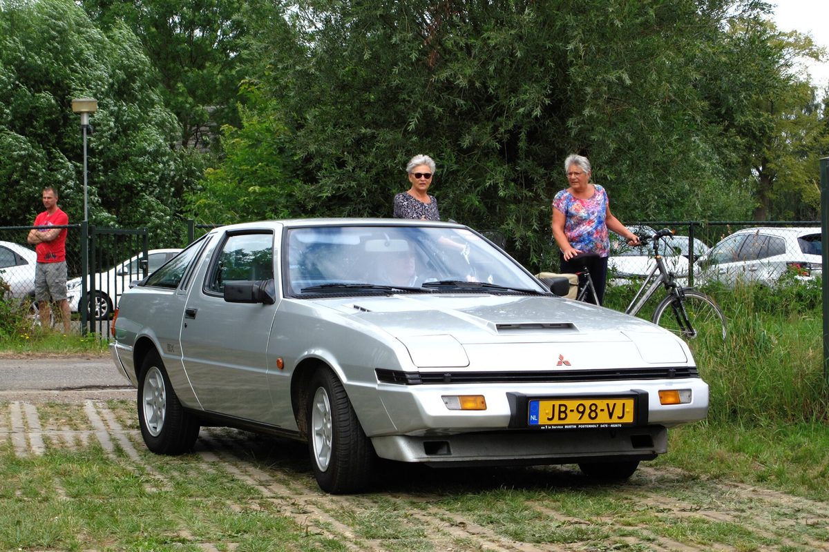 A silver Mitsubishi Starion hatchback coupe parked on grass at a classic car meet, with onlookers behind it