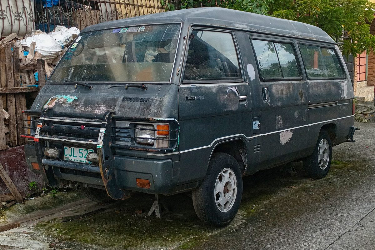A weathered dark gray Mitsubishi L300 van with peeling paint and a front brush guard, parked on a mossy street