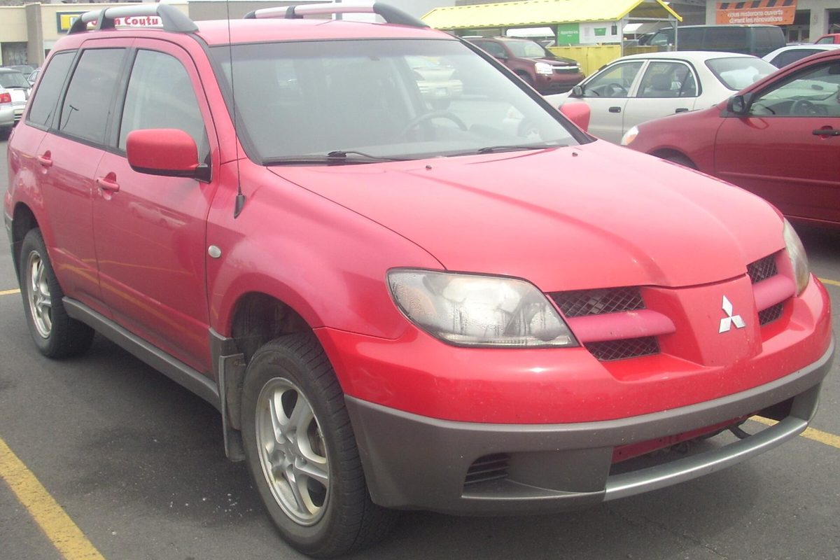 A red first-generation Mitsubishi Outlander SUV with roof rails and a gray lower bumper, parked in a store lot with other cars behind it