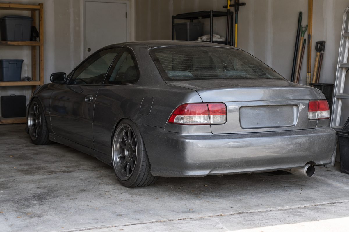 A lowered gray two-door coupe on dark aftermarket wheels parked in a garage beside storage shelves, a ladder and yard tools