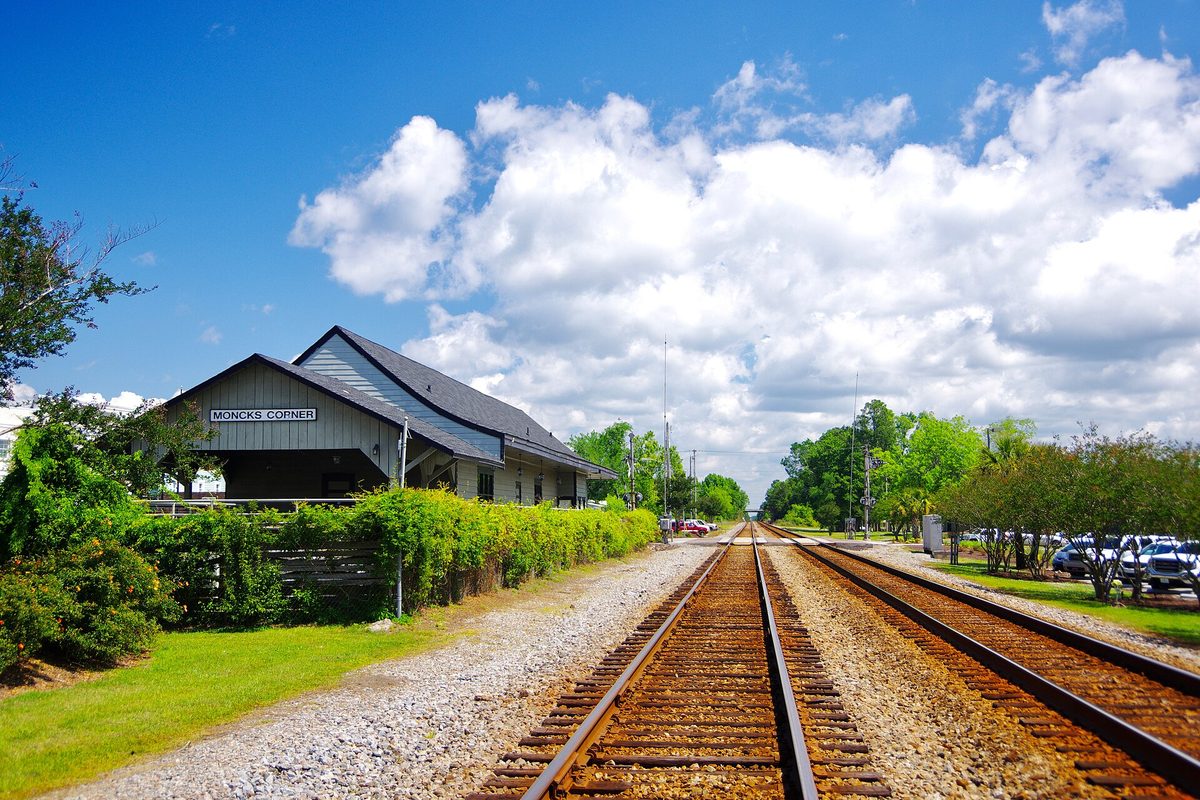 The gray wooden Moncks Corner train depot with its name sign, beside a pair of railroad tracks under a blue sky with white clouds