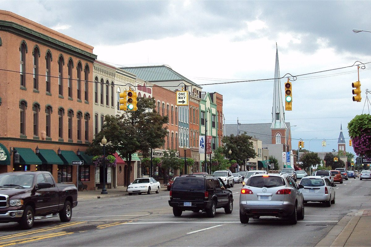 A downtown Monroe street lined with brick storefronts and green awnings, cars waiting at a traffic light and a white church steeple behind