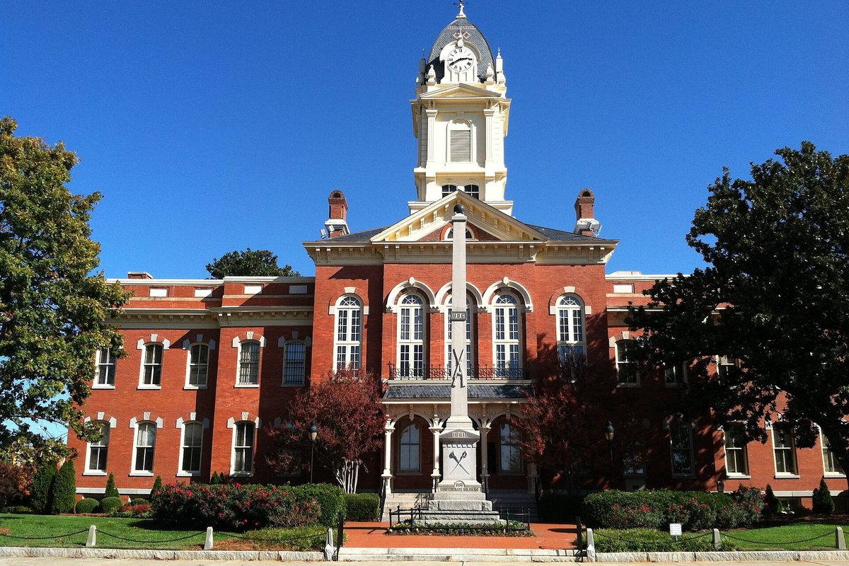 Monroe's red brick historic courthouse with a white clock cupola and a tall stone monument in front, under a clear blue sky