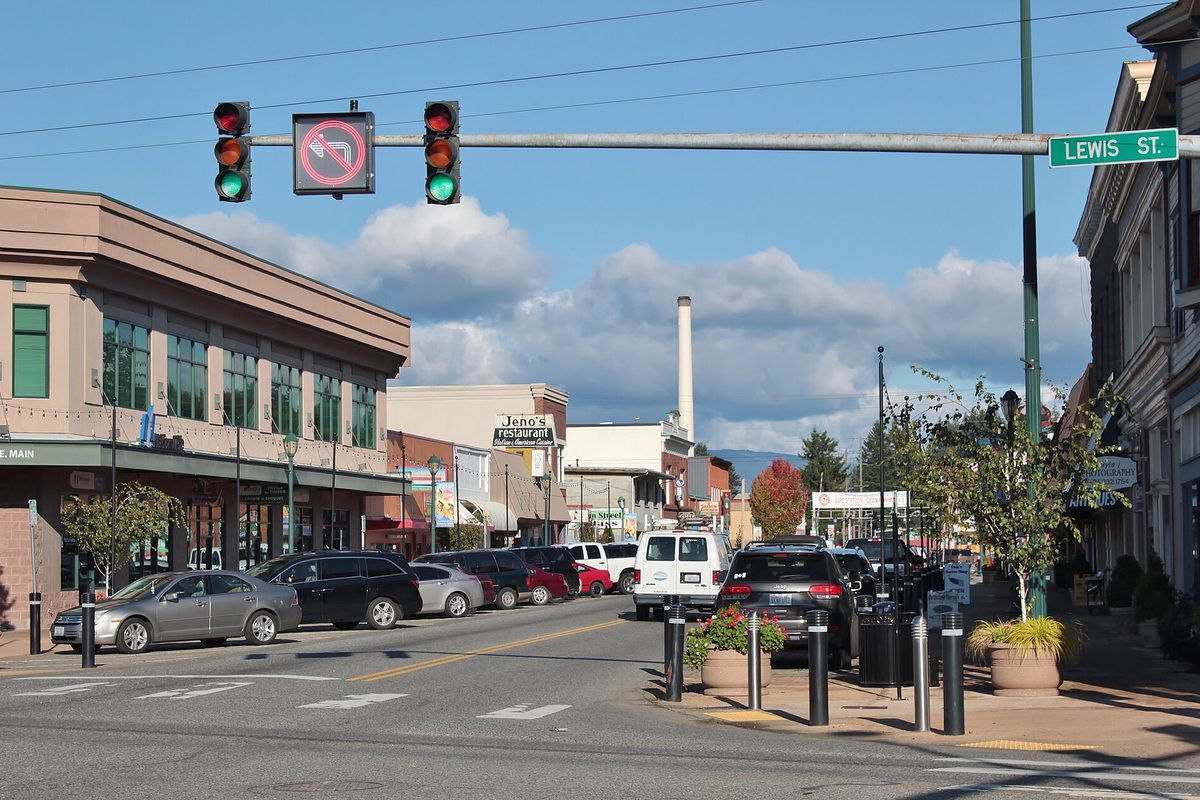 Traffic lights over the Main Street and Lewis Street intersection in downtown Monroe, with parked cars, storefronts and a tall smokestack under a blue sky
