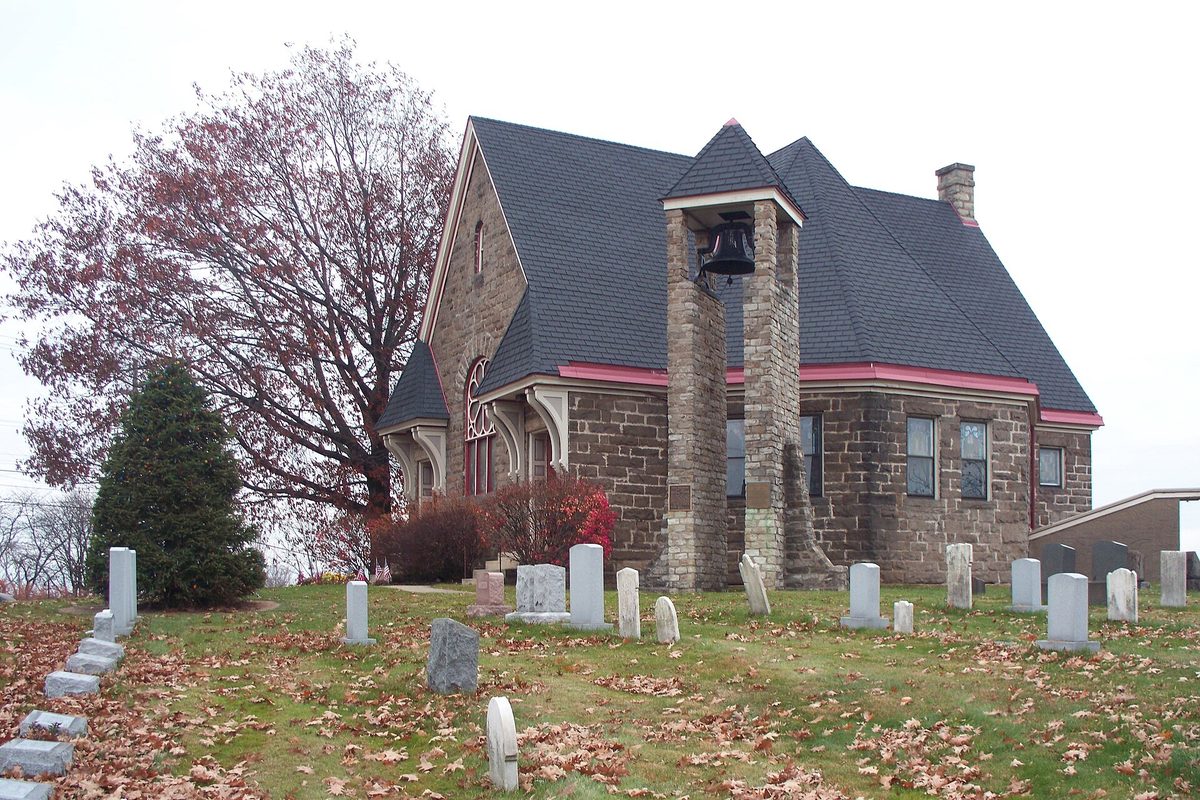 The Old Stone Church in Monroeville, a stone building with a gray roof and a bell hung in an open stone tower, old gravestones on a leaf-covered lawn in front