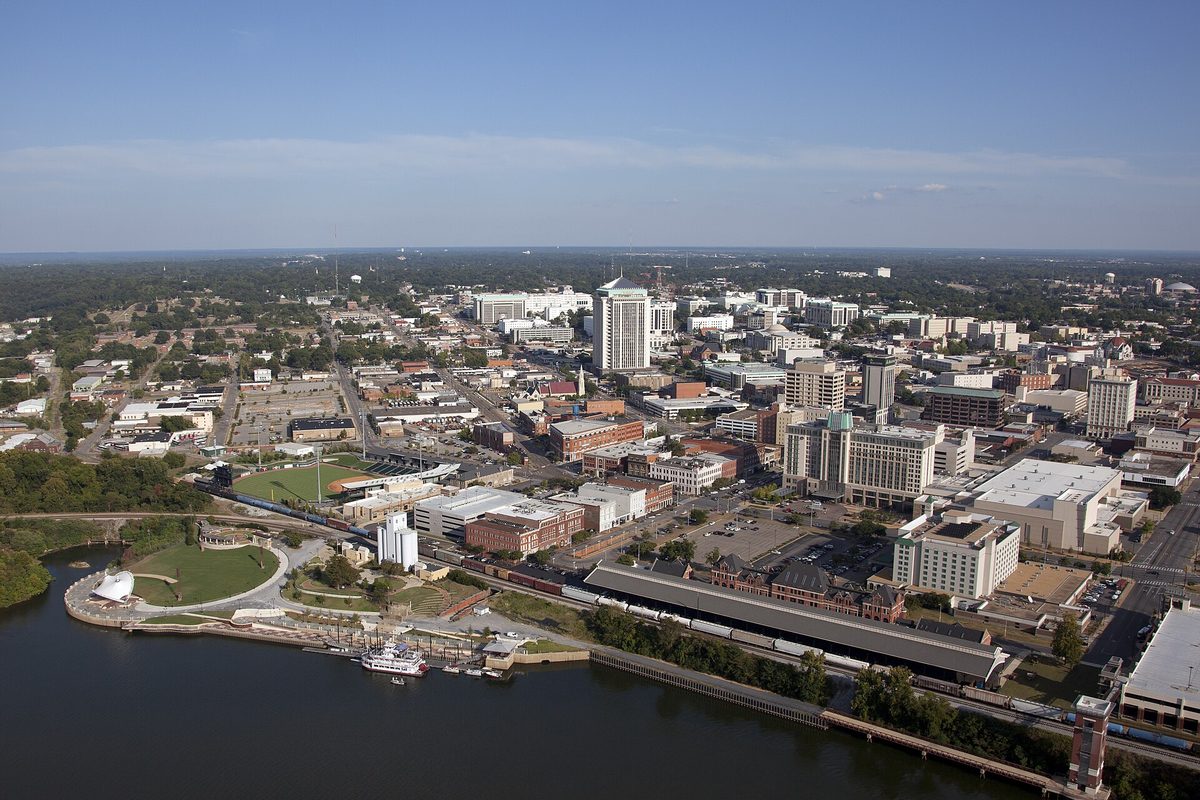 Aerial view of downtown Montgomery beside the river, with a riverfront amphitheater, a moored paddle-wheel boat and a long rail shed along the water