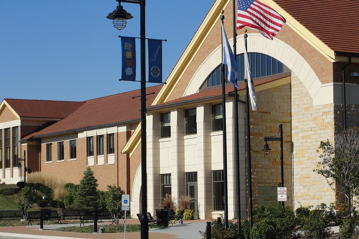 Montgomery Village Hall, a tan stone and brick building with a large arched window and red tile roof, with flags and a banner-hung lamp post out front