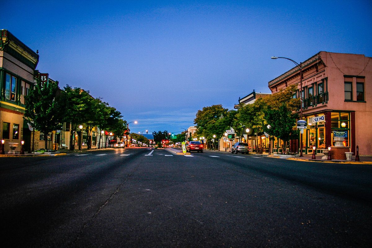 Downtown Montrose at dusk, an empty main street lined with lit storefronts, street lamps and trees under a deep blue sky