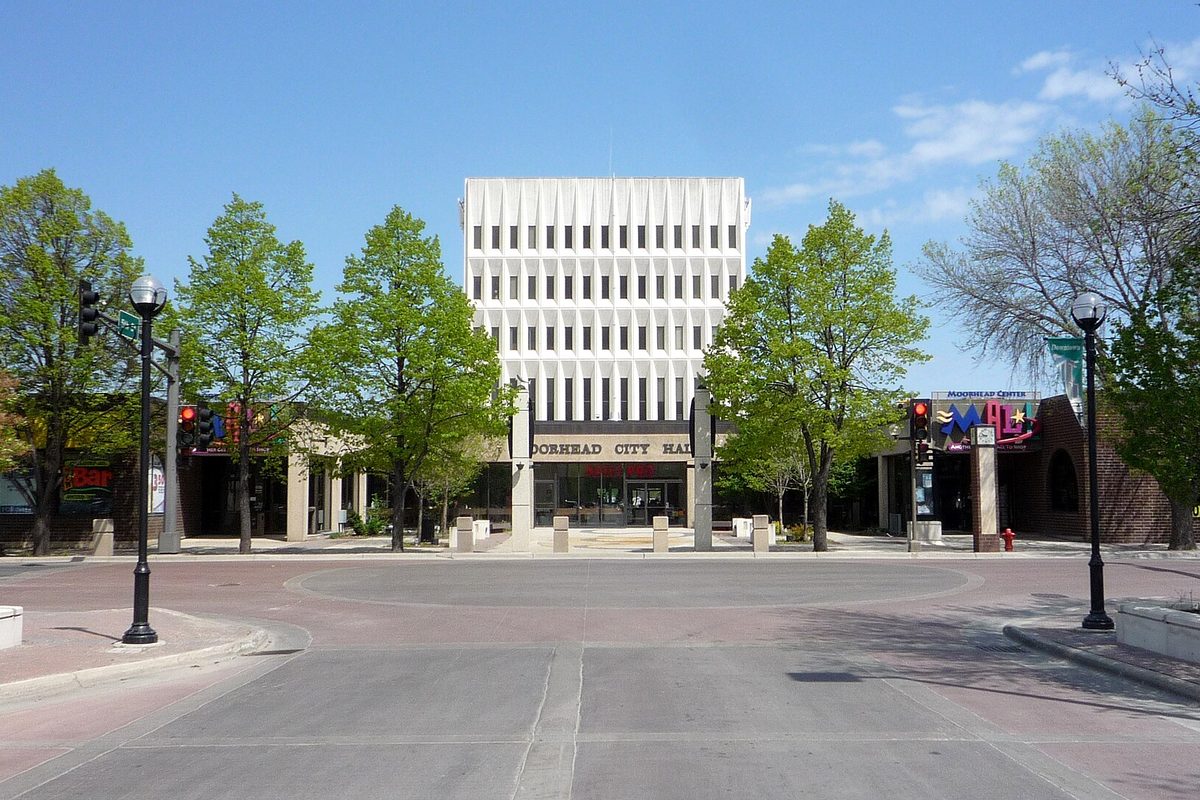Moorhead City Hall, a tall white building with narrow vertical windows, at the end of a brick plaza lined with green trees and street lamps