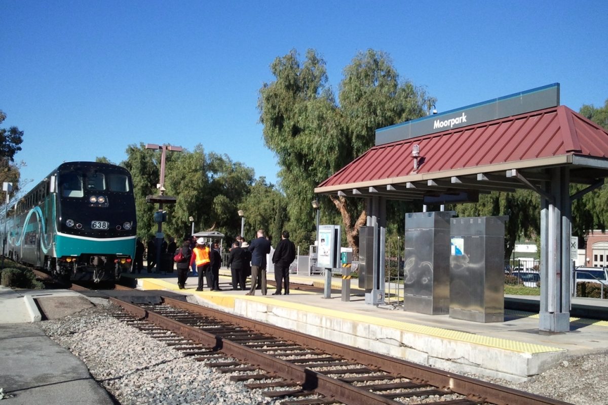 A Metrolink train stopped at the Moorpark station platform beside a red-roofed shelter with a Moorpark sign, under a clear blue sky