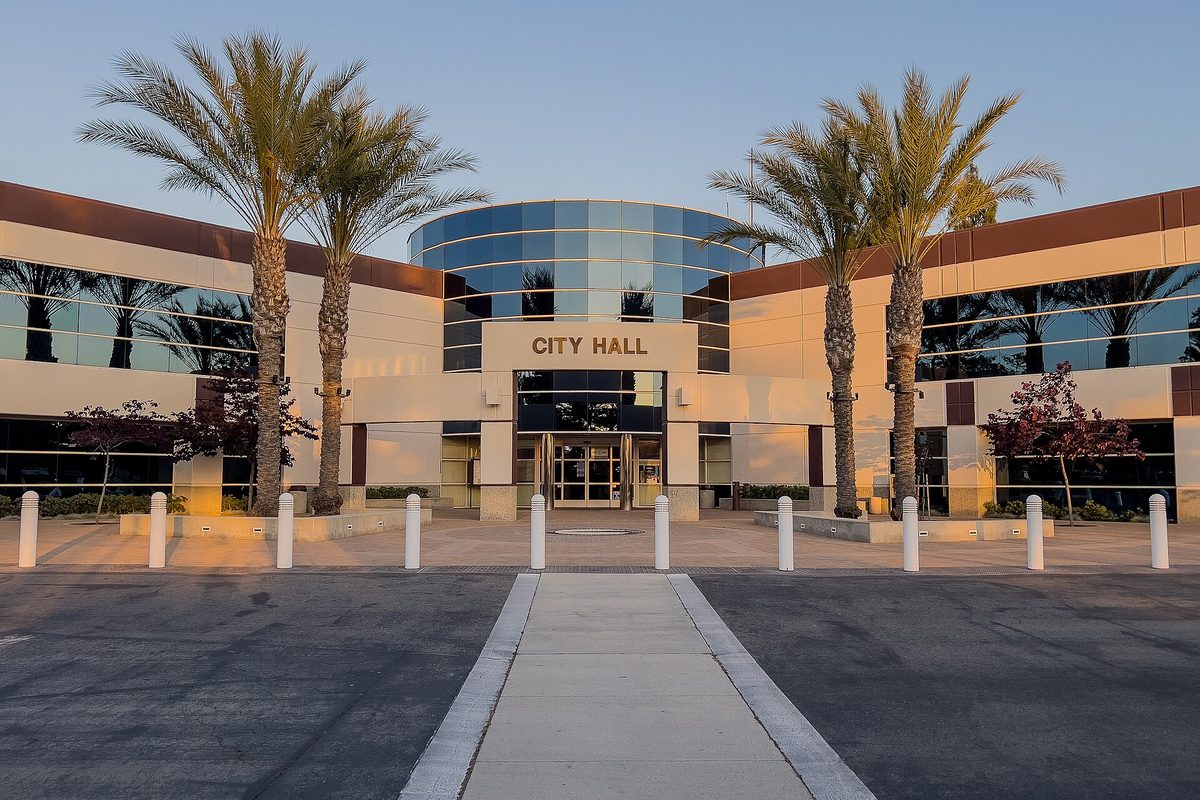 Moreno Valley City Hall at dusk, a tan building with a curved blue glass entrance and City Hall lettering, flanked by palm trees, with white bollards along the walkway