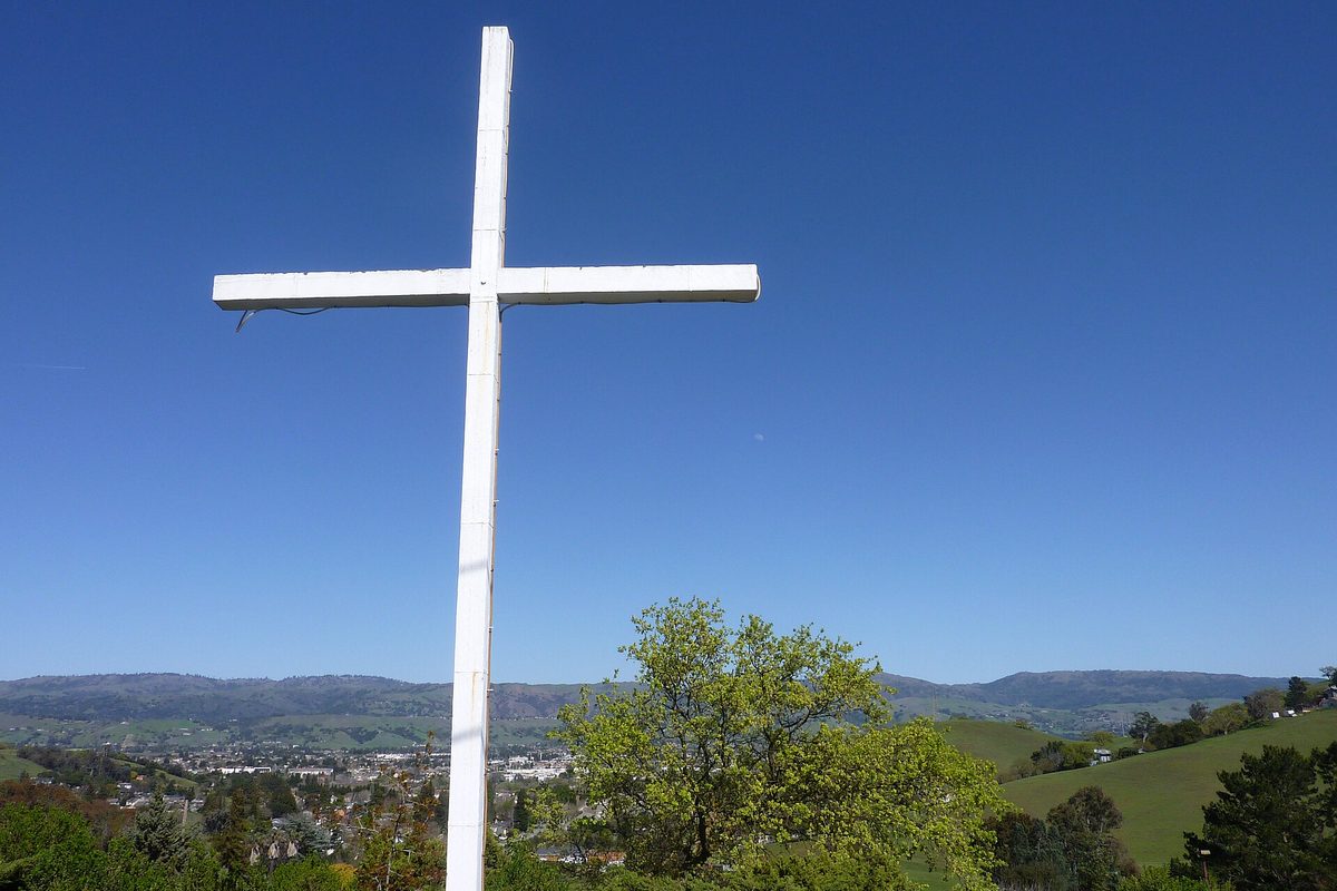 A tall white cross on a green hillside, with the town of Morgan Hill and a ridge of hills below under a clear blue sky