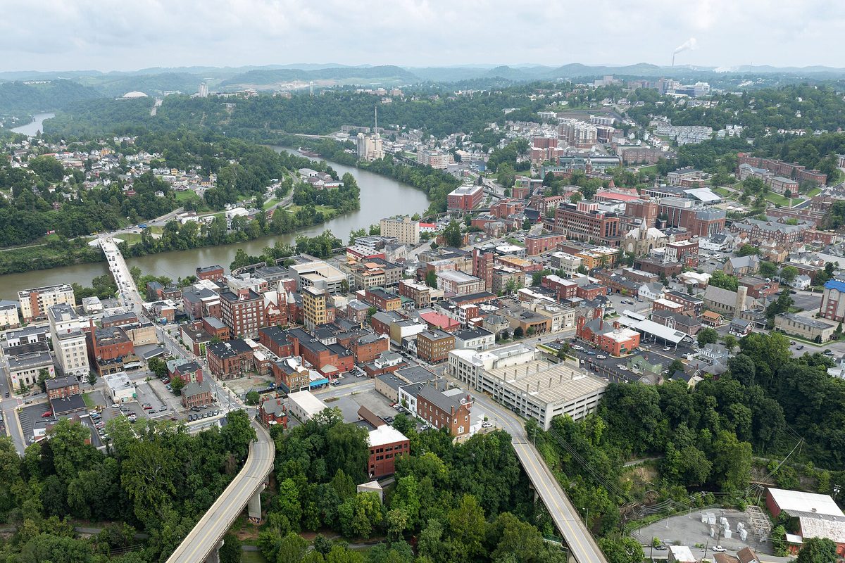 Aerial view of Morgantown's brick downtown beside the curving Monongahela River, with an elevated concrete guideway in the foreground and green hills beyond