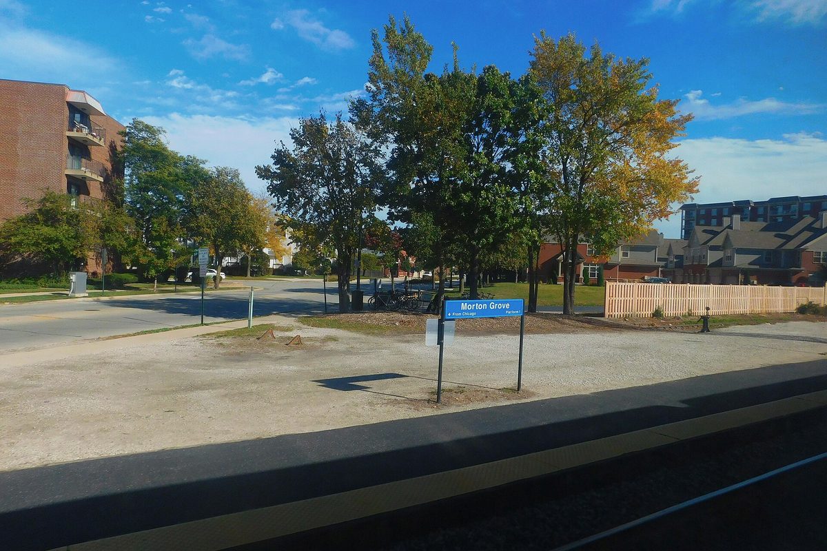 A blue Morton Grove station sign beside the Metra tracks, with trees, a gravel lot and apartment buildings behind it, seen from a passing train